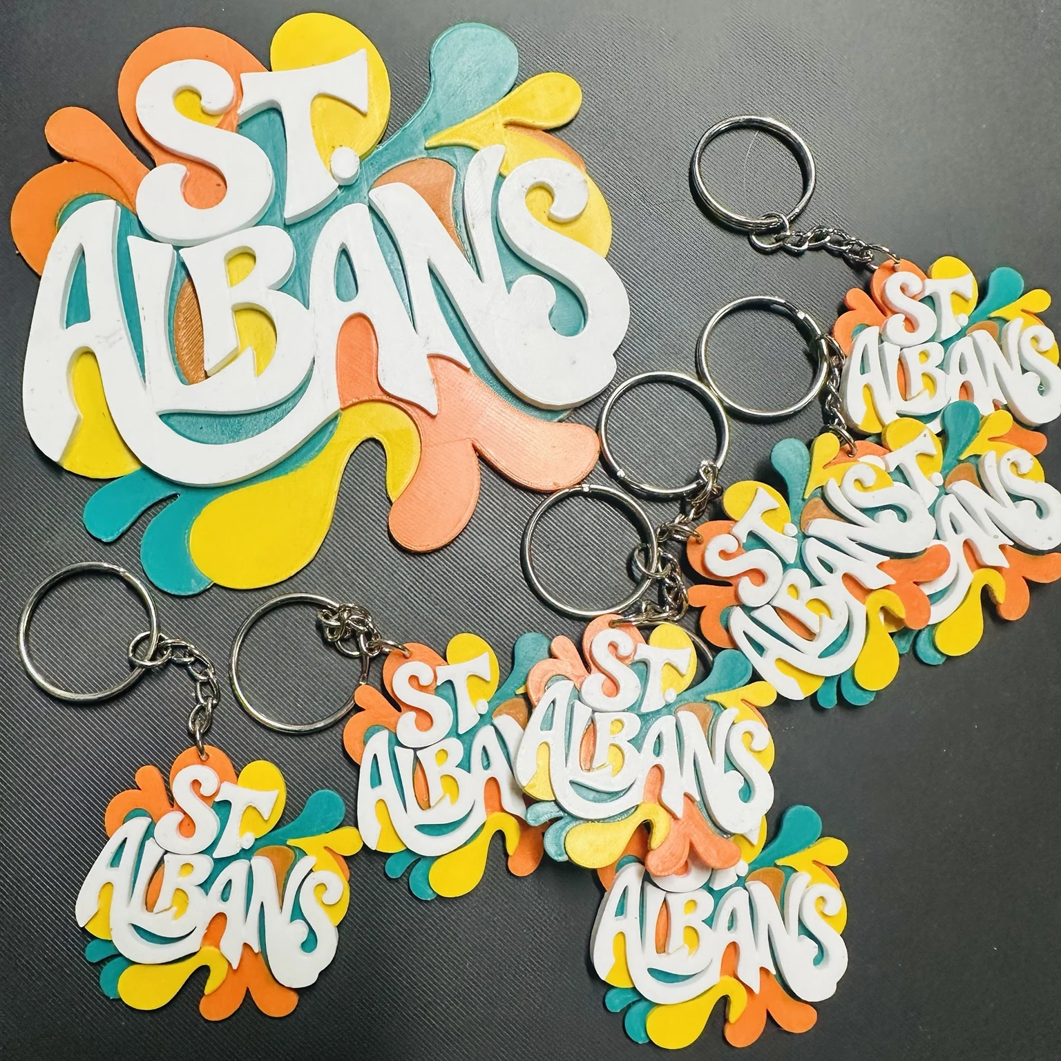 Custom St. Albans keychains and a larger printed logo with raised white lettering over teal, orange, and yellow shapes
